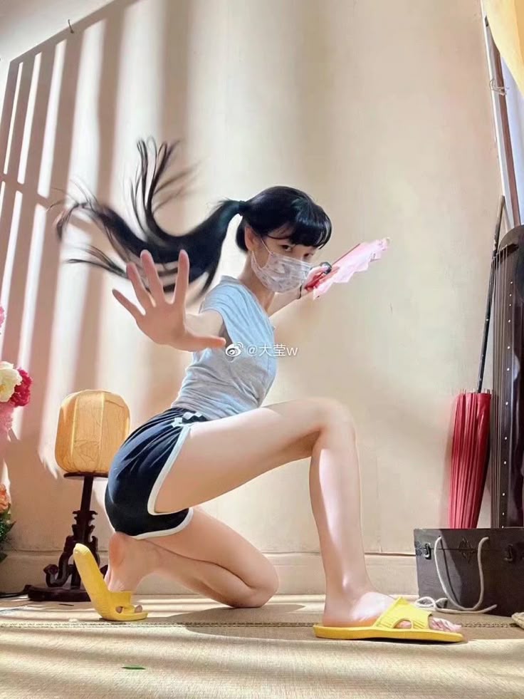 a young woman wearing a grey tshirt, black shorts with white trim, a white face mask, and yellow sandals is kneeling and holding her hand up in a gesture. her black hair is styled in a ponytail, and she has bangs. she is holding a pink fan in her left hand. behind her, there is a traditional musical instrument, a red umbrella, and a small table lamp. the background is a beige wall.