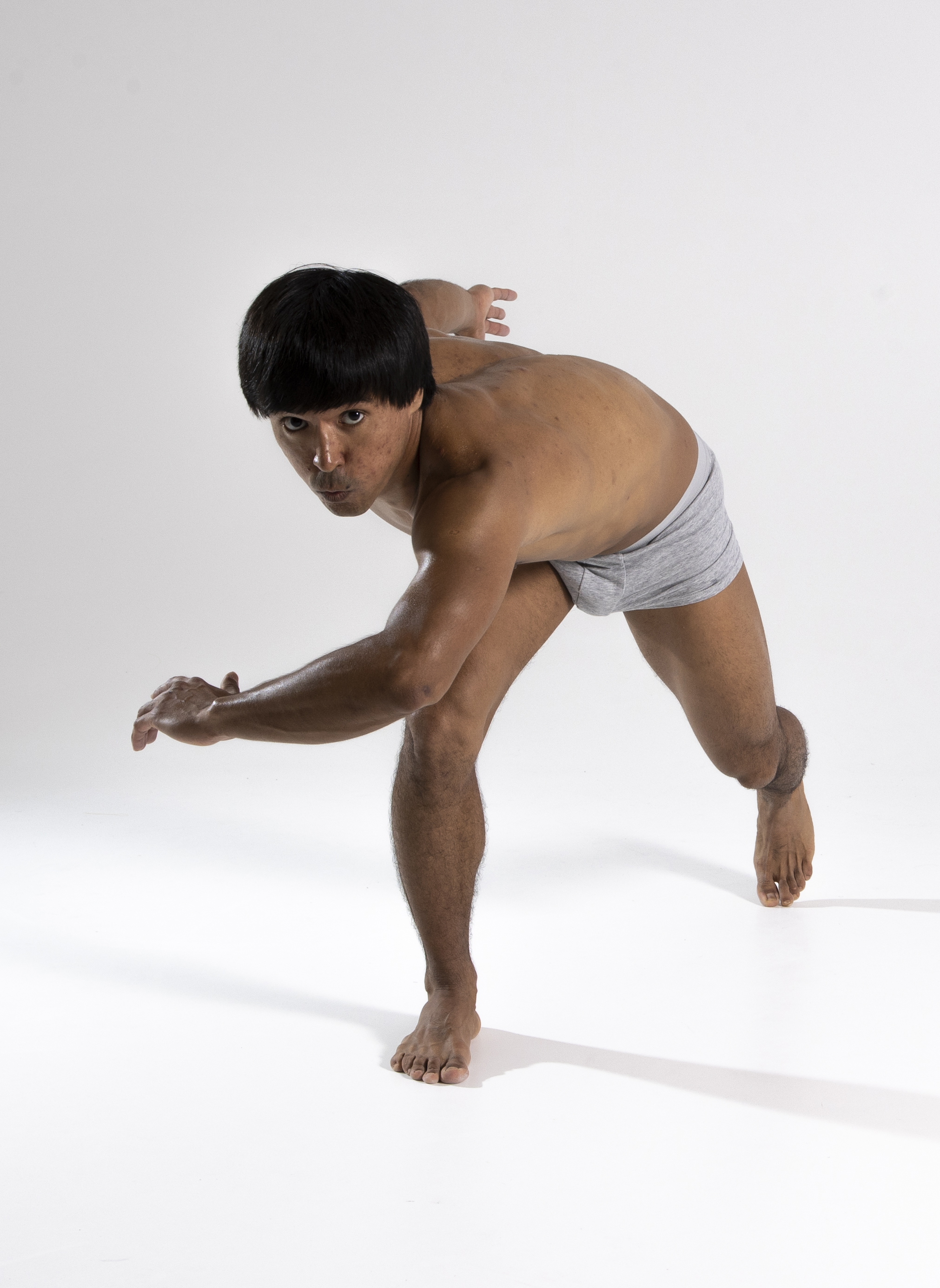 a shirtless man with a bowl cut and wearing briefs is barefoot and performing a yoga pose or a contemporary dance move in a studio against a white background. he's looking directly at the camera with a focused expression.