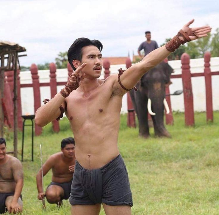 a shirtless, muscular man with a serious expression is standing in a field,  with his arms outstretched as if he is throwing a spear. he is wearing a pair of dark shorts and traditional thai wristbands. the man's torso is on full display, showcasing his muscular physique. the background features a wooden fence and a large elephant standing in the distance. the setting is outdoors and the grass is lush and green, suggesting a rural or historical location. this photo captures the athleticism, tradition, and power associated with the man.