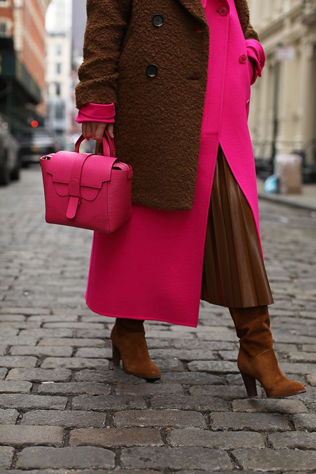 a stylish woman strolls down a cobblestone street in new york city.  she's wearing a brown fluffy coat over a vibrant pink coat with a brown pleated leather skirt.  she carries a bright pink senreve handbag and wears brown suede heeled boots. the overall look is chic and autumn/winter appropriate. the image is wellcomposed, focusing on the woman's outfit and accessories.