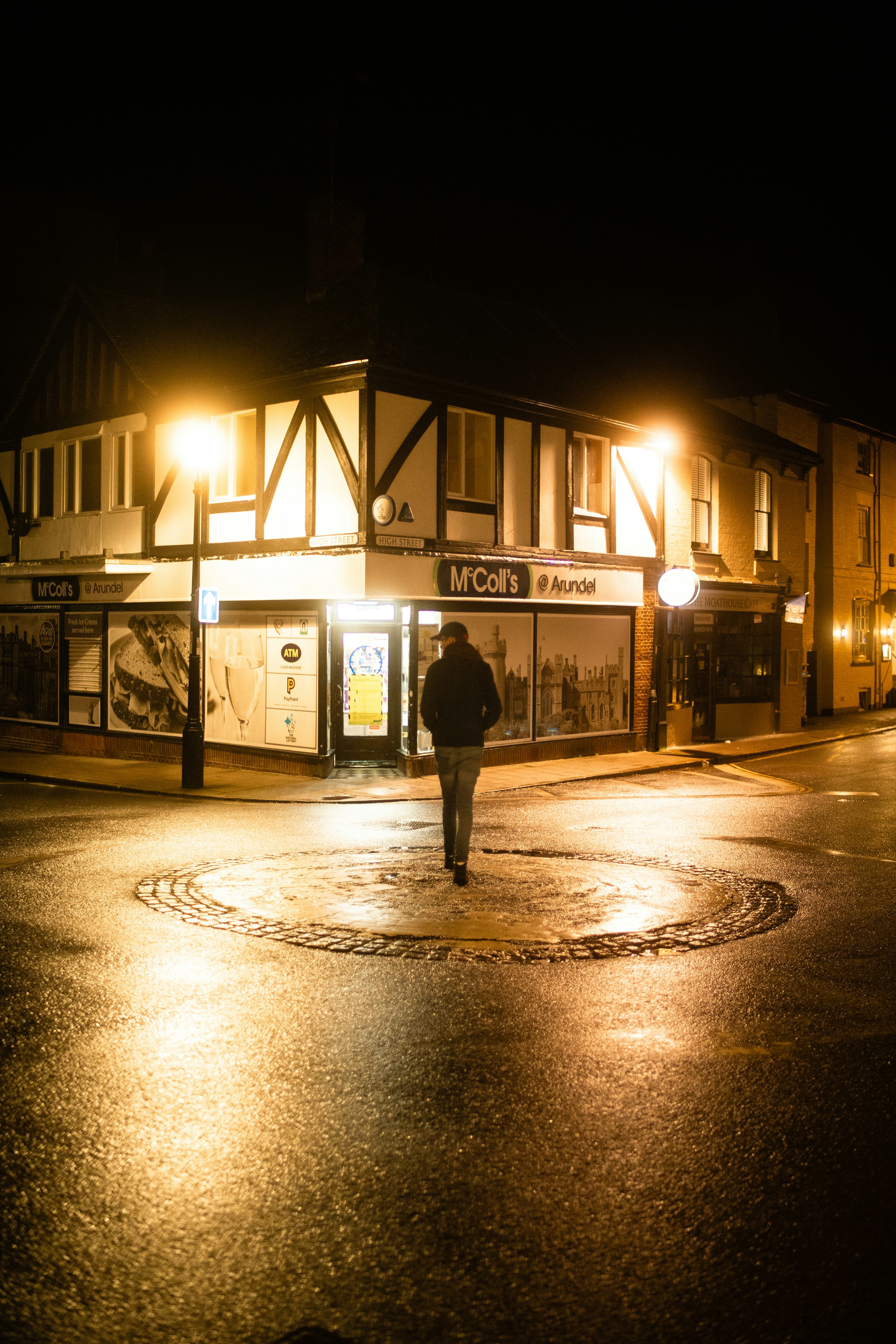 a man walks through a puddle on a street at night. the mccoll's convenience store is located on the corner of the street.