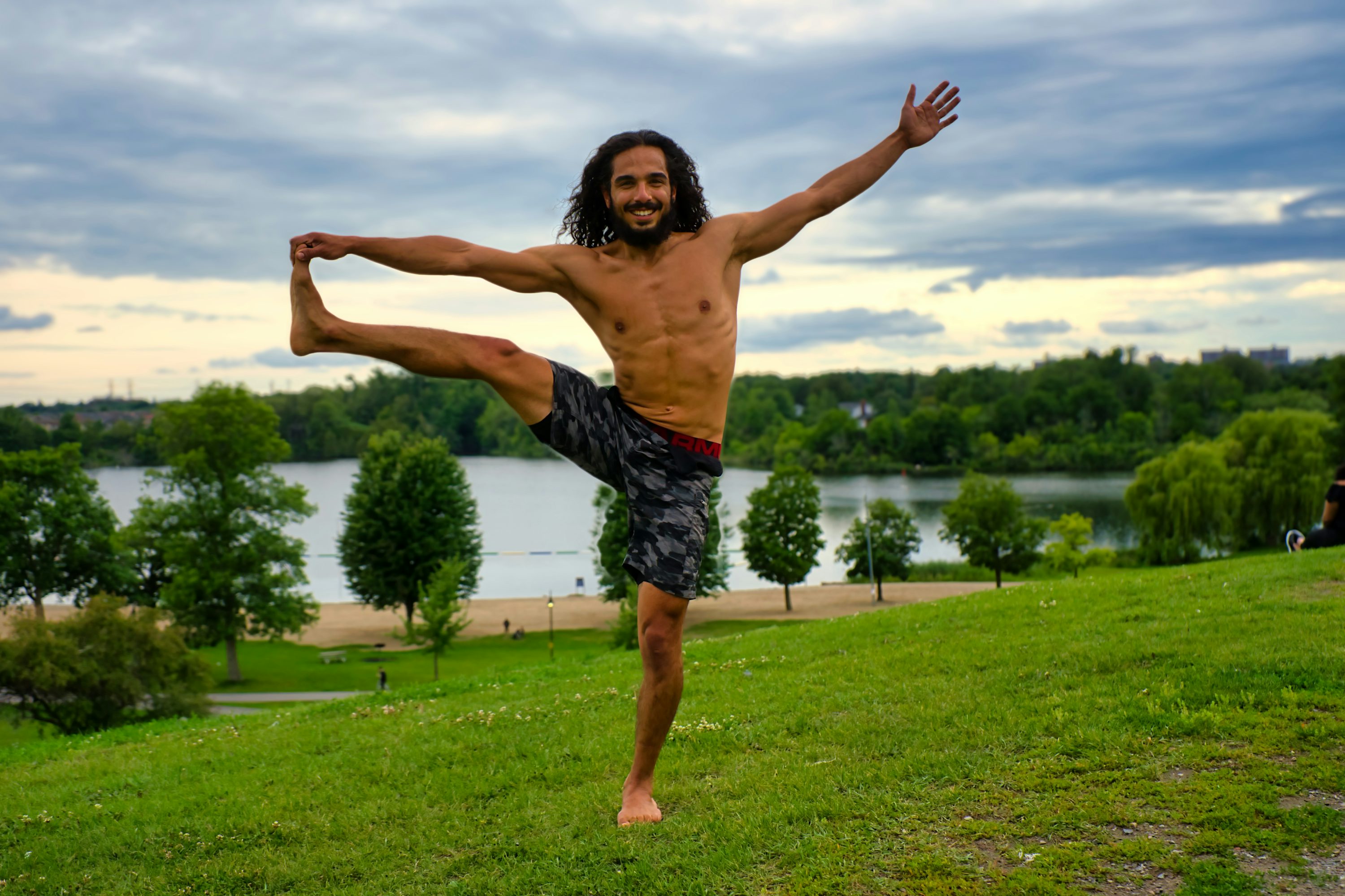 a fit man with long brown hair is performing a yoga pose outdoors in a park. he is barefoot, wearing athletic shorts, and has his leg raised high in the air. he is smiling and looks happy and peaceful. the background is a beautiful lake and park scenery with green grass, trees, and a sunset sky. the overall feeling is one of serenity, health, and wellbeing.