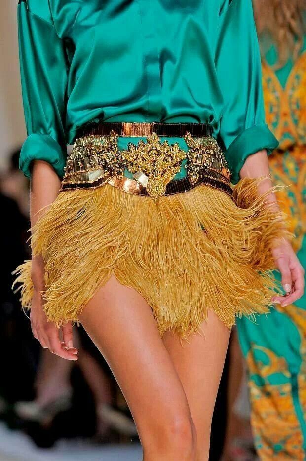 a model is shown on a runway, wearing a vibrant green satin blouse paired with a short, mustardyellow fringe skirt. the skirt is cinched at the waist with a heavily embellished, broad belt featuring metallic accents and ornate detailing. the model's legs are visible as she stands, presenting the ensemble. in the background, other individuals and elements of the runway setting are partially visible.