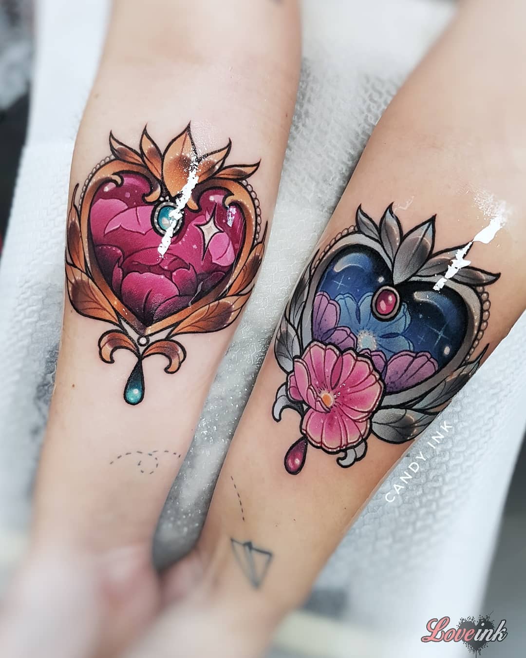 the image shows two forearms with matching tattoos. the tattoos are of ornate heartshaped lockets with jewels and flowers. the locket on the left is gold with a pink jewel and a pink teardrop gem dangling from the bottom. the locket on the right is silver with a blue and purple galaxy design and a pink flower at the bottom. the tattoos are done in a neotraditional style with bold lines and bright colors. the tattoos are by the artist "candy ink".