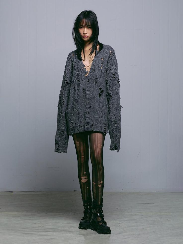 a fullbody shot of a young asian woman posing in a studio setting against a light grey background. she is wearing an oversized grey distressed cardigan with multiple rips and tears throughout, paired with black fishnet tights that also have rips and tears, and chunky black combat boots. her hair is dark and straight, and she has minimal makeup. the overall style is edgy and grunge. the focus is on the model and her clothing, showcasing a fashionforward look.