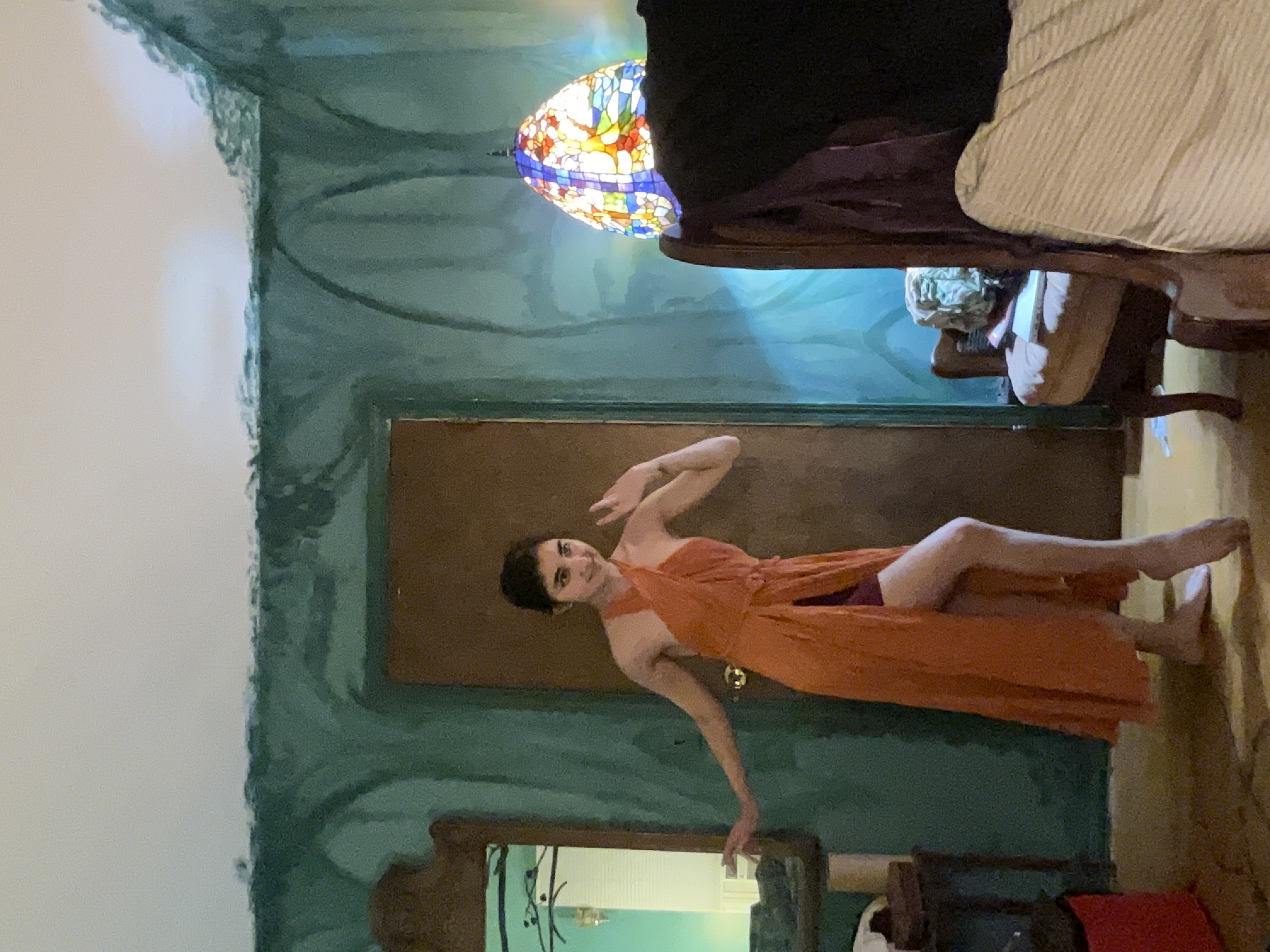 a young woman in a burnt orange halter wrap dress with a high slit stands in a room with teal walls. she poses with one hand on her hip. the room features vintage furniture including a highbacked chair and a stainedglass lamp. the overall aesthetic is bohemian or eclectic. the image appears to be a photograph.
