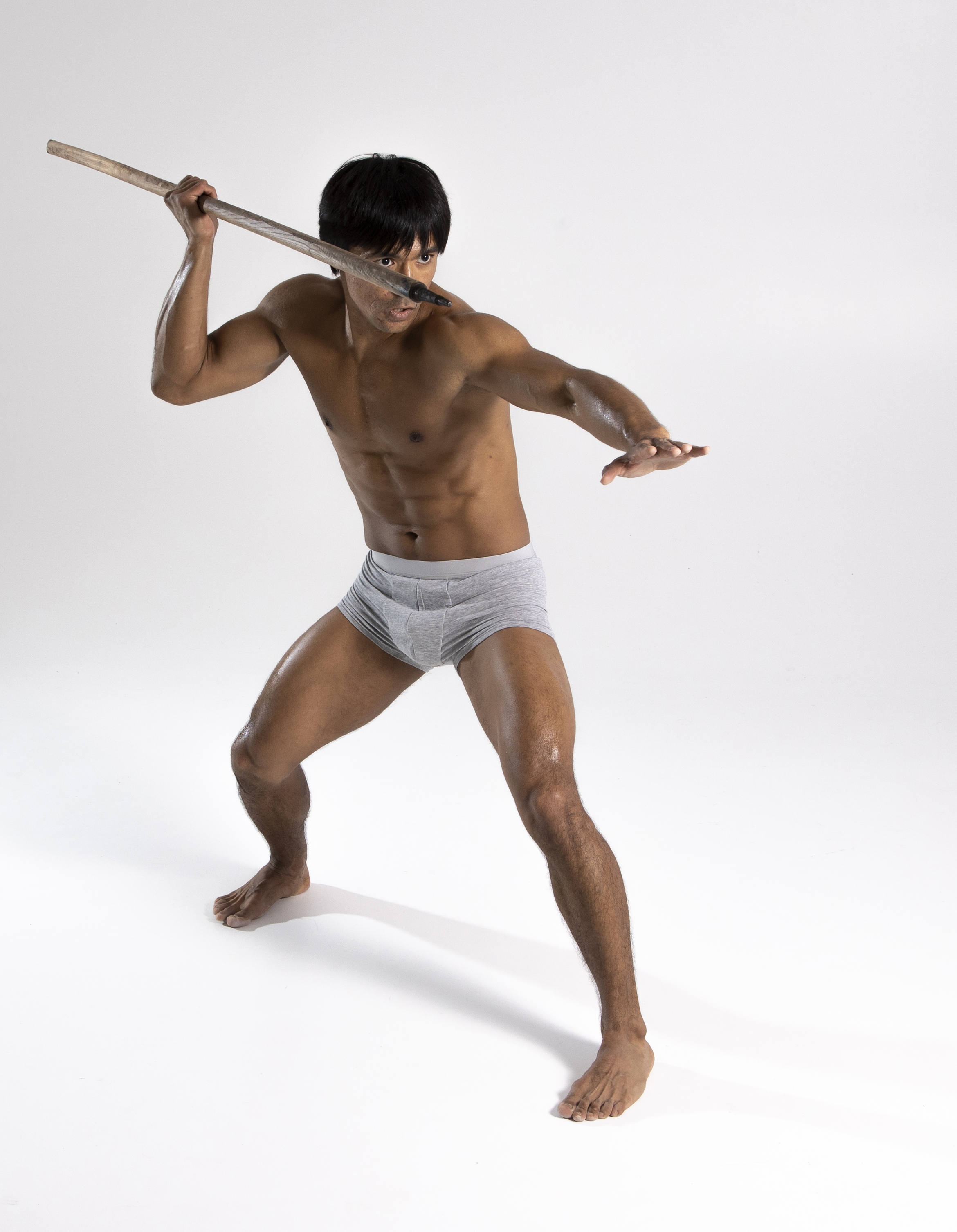 a shirtless man in gray underwear stands on a white background, holding a wooden spear. he is in a poised, athletic stance, ready to throw the spear. he has a toned physique and is barefoot. the photograph is a full body studio shot.