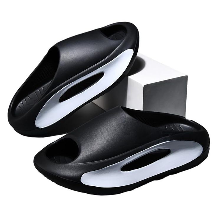 two black and silver slides are displayed against a white background with a geometric white object behind the top shoe. the slides have a modern design with a cutout section and a sleek, comfortable look.