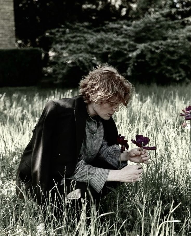 a young, redhead male model is captured in a black and white photo, crouched in a field of wildflowers. he is wearing a dark jacket and shirt, and he is holding a black flower in his hand. the background of the image is a lush forest, creating a serene and natural setting.  the photo evokes a sense of tranquility and connection with nature.