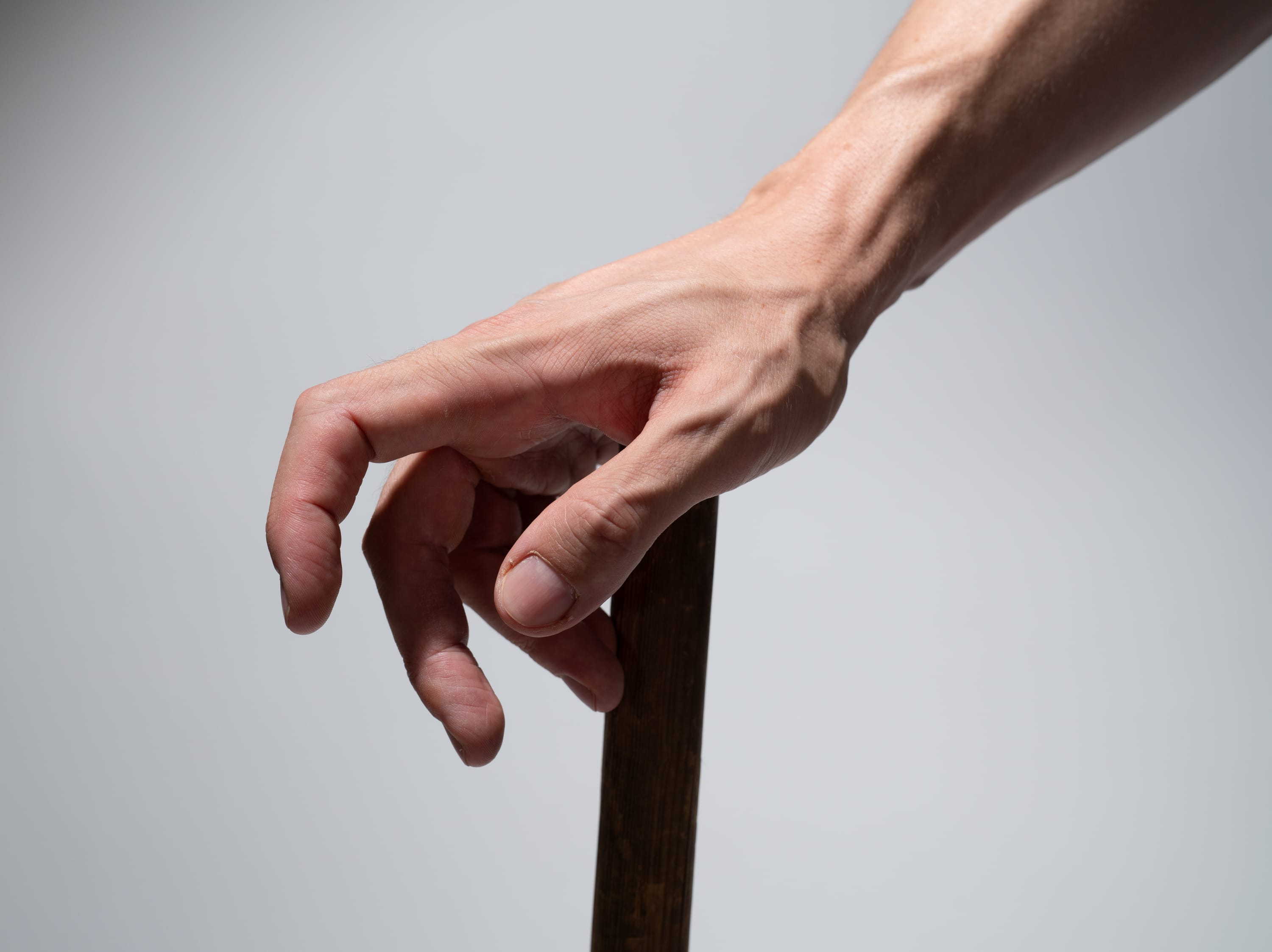 a closeup view of a man's hand gripping a wooden walking stick. the hand exhibits prominent veins and a firm grasp, conveying a sense of strength and reliance on the cane for support. the neutral background and studio lighting emphasize the hand and the walking stick as the central focus.