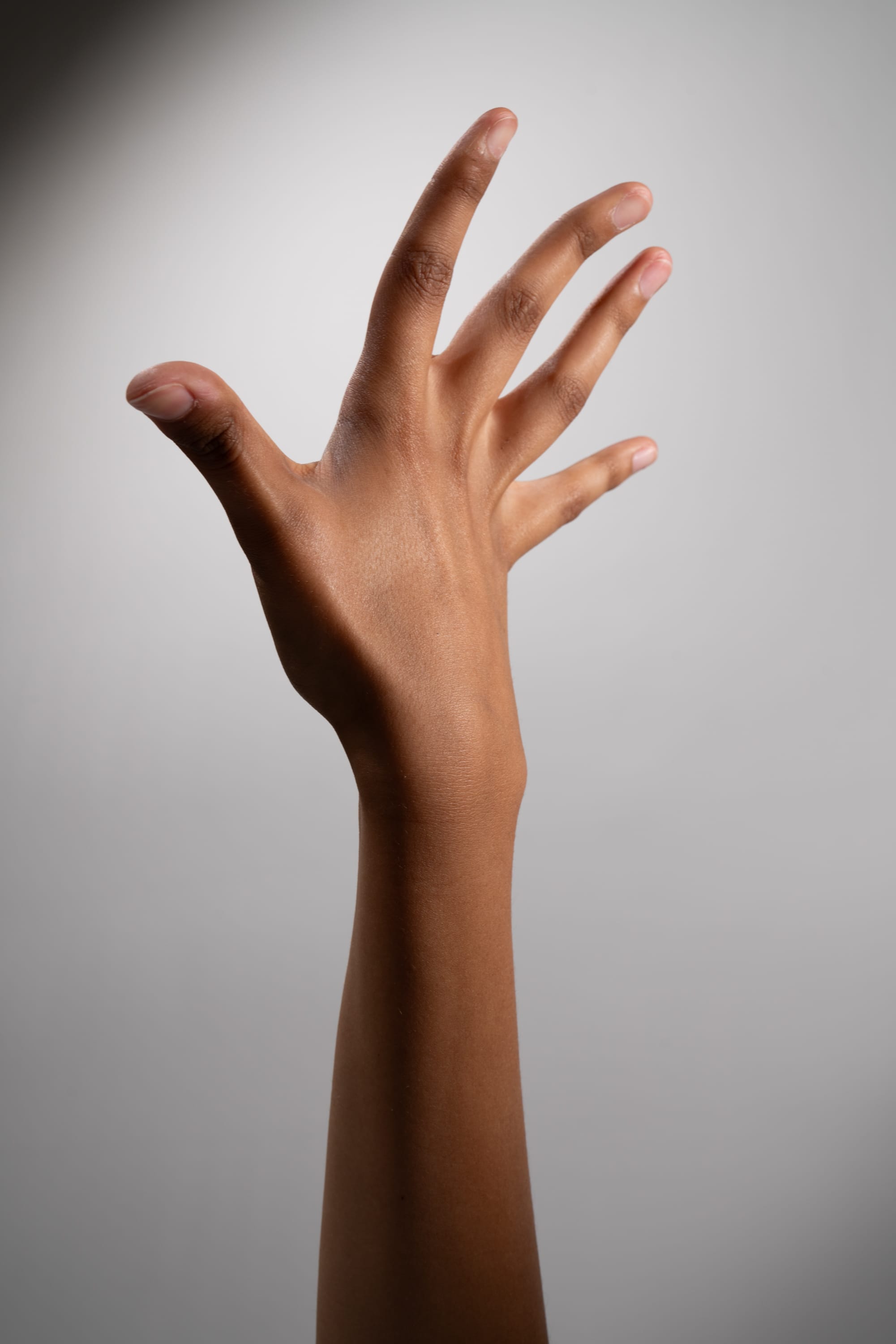 an image of a woman's arm and hand extended upwards against a gray background. the hand is open, with the fingers spread apart, and the arm is slender and graceful. the lighting is soft and directional, creating a sense of depth and dimension.
