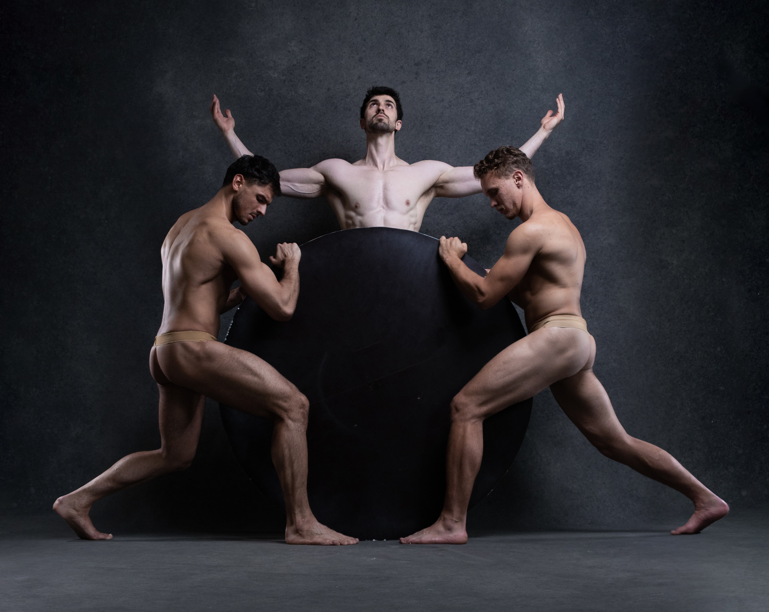 the photograph showcases three muscular male dancers in a striking pose. against a dark, textured studio backdrop, two of the dancers flank a large, black circular prop. they grip its edges, their bodies taut with effort. their gazes are fixed downward, adding to the image's intensity. in the center, a third dancer stands with arms outstretched, his body framed by the others. the dramatic lighting accentuates their physiques and the raw energy of their movements.  the overall composition evokes themes of strength, balance, and the human form in motion.