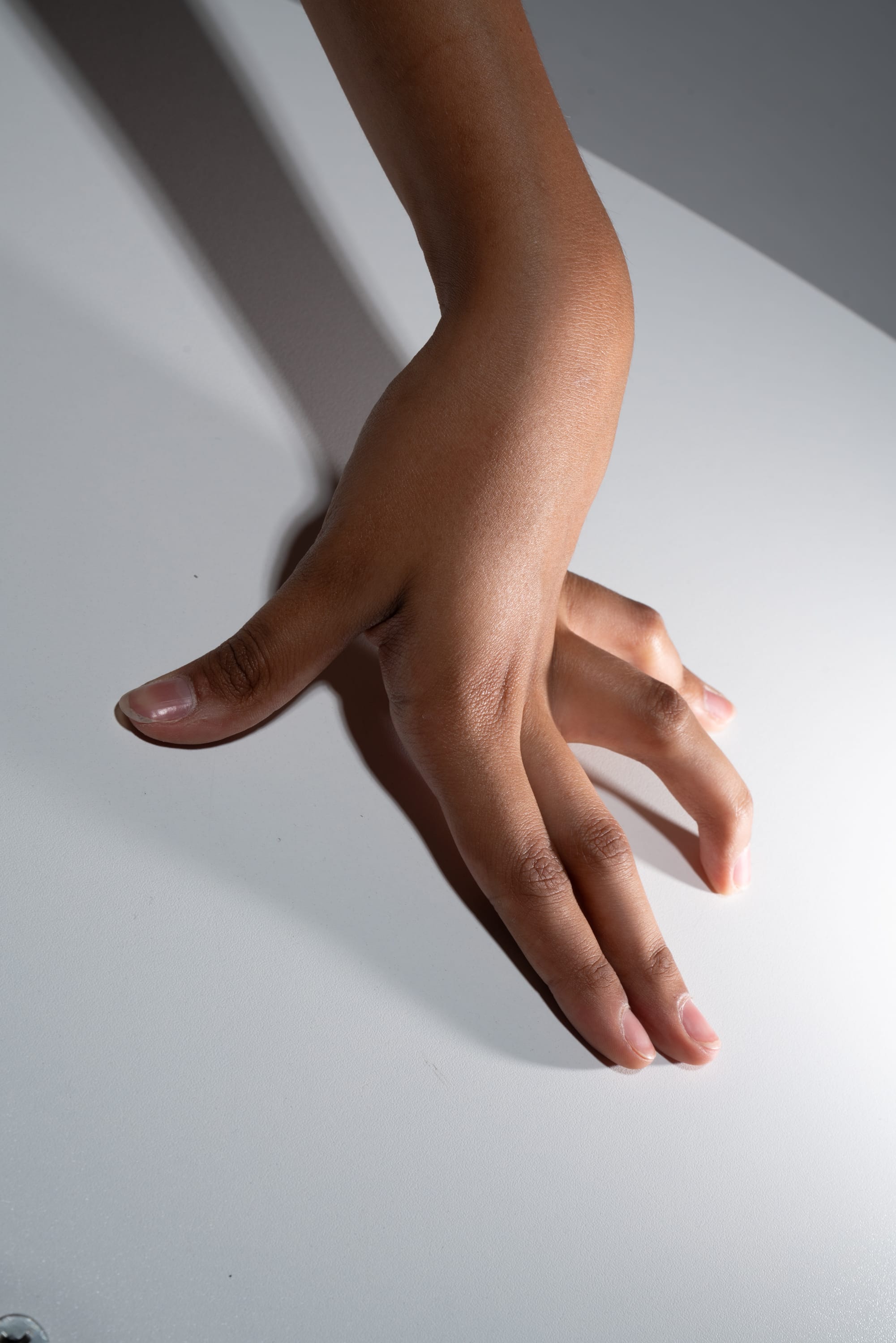 a closeup photograph of a hand with dark skin gently splayed on a smooth, white surface. the image is lit with artificial light, creating dramatic shadows and emphasizing the hand's texture and form.  the highangle perspective and minimalist aesthetic draw attention to the detail and subtle gesture of the hand.