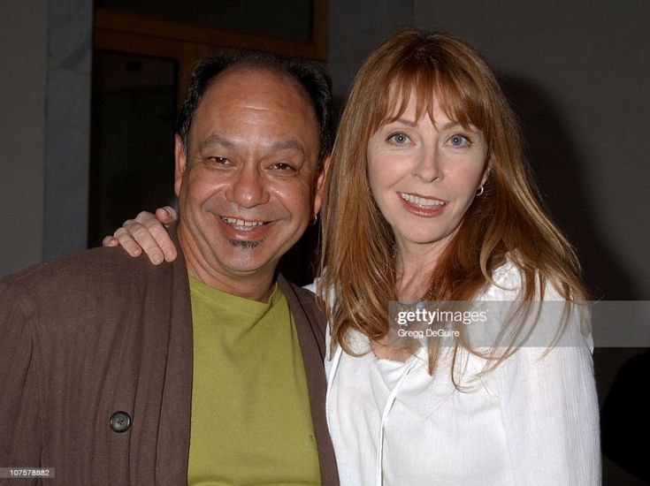 a man and woman, dressed up and smiling, are posing for a photo, looking happy and comfortable together.  the man is wearing a brown jacket over a green shirt and the woman is wearing a white buttondown shirt. they are standing close together in a casual pose, possibly at an event or a party. the man is putting his arm around the woman.