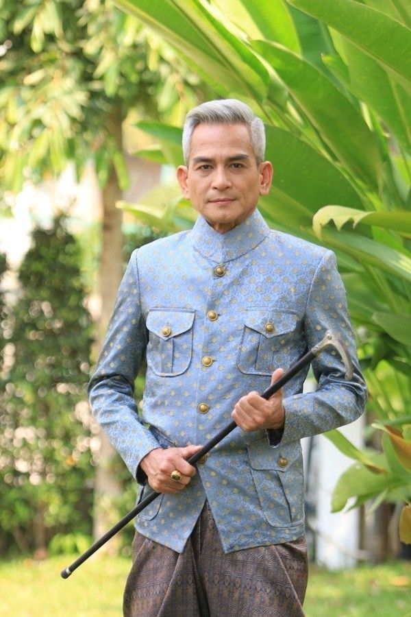 a man in traditional thai clothing is standing outdoors in a garden setting, with a green leafy background. he is smiling and holding a walking stick. he is wearing a light blue patterned jacket and a brown patterned skirt with gold buttons on the jacket.