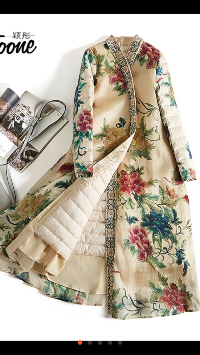 a long, floral patterned coat is displayed. the coat features an intricate floral design with a beige base color and various hues of red, pink, blue, and green in the floral elements. it has a quilted lining in a light beige color, visible where the coat is slightly open. an ornate border runs along the edges of the coat. beside the coat is a book and accessories.