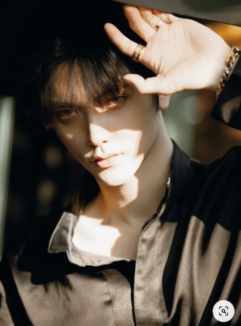 he image is a closeup portrait of a young man with dark hair and sharp features. he is wearing a dark, silky shirt, unbuttoned to reveal a plain undershirt. he is accessorized with multiple rings on his fingers and a bracelet on his wrist.  the lighting is dramatic, with sunlight streaming in from the side, casting strong shadows across his face and body.  he is shielding his eyes from the sun with his hand, creating a sense of mystery and allure.  the overall mood of the image is stylish and edgy.