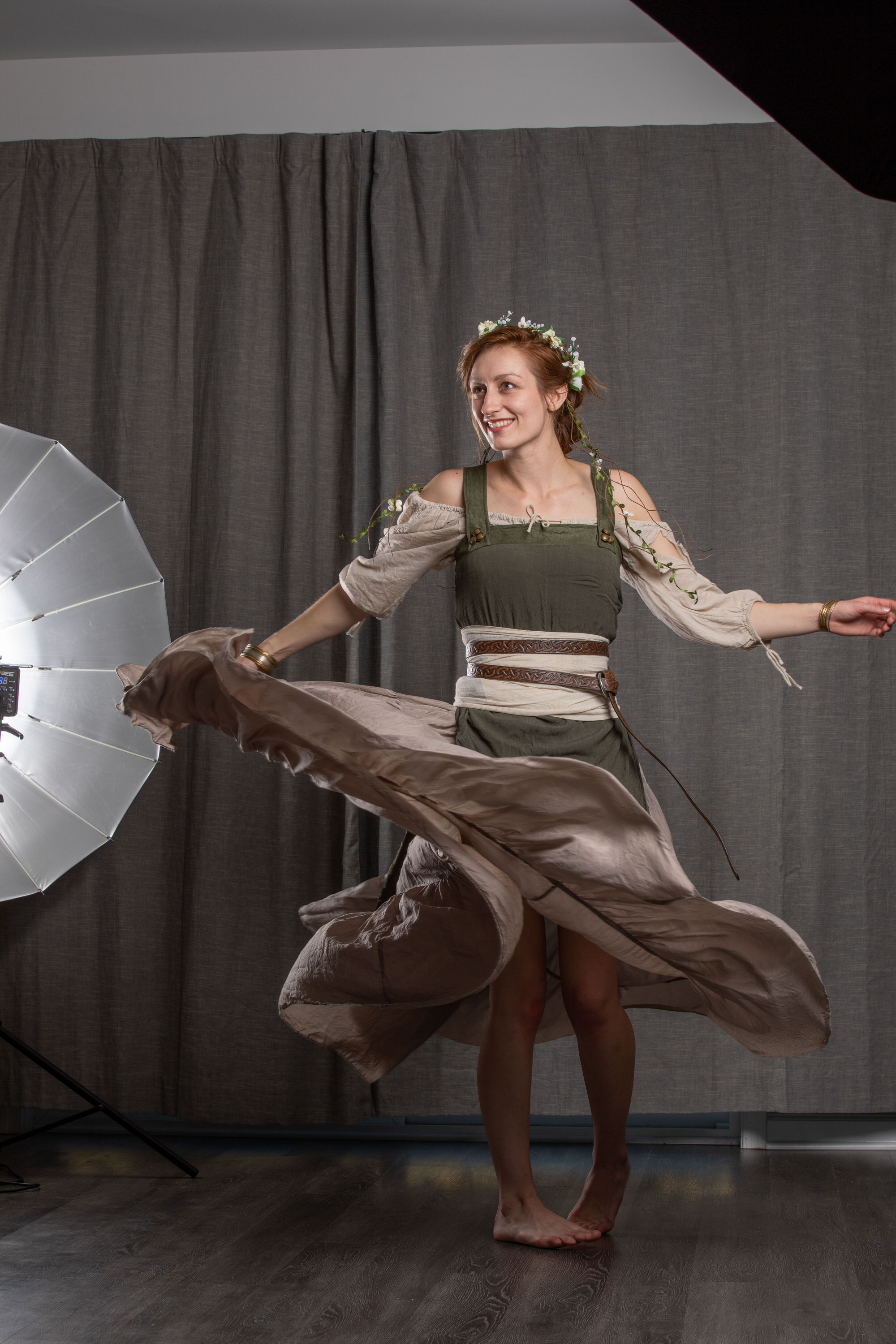 a woman in a medievalinspired dress twirls in a photography studio. she is barefoot and smiling, her dress flowing around her. a large softbox illuminates her from the left, casting a soft light on the scene.  she has reddish hair and is wearing a floral crown. the dress is a light brown with a green bodice. the background is a simple grey curtain.