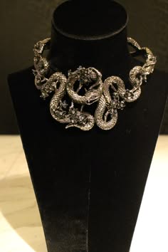 a closeup of a silver snake necklace on a black mannequin. the necklace is ornate and detailed, with intricate snake scales and flowers. the necklace is a statement piece that would add a touch of glamor to any outfit.