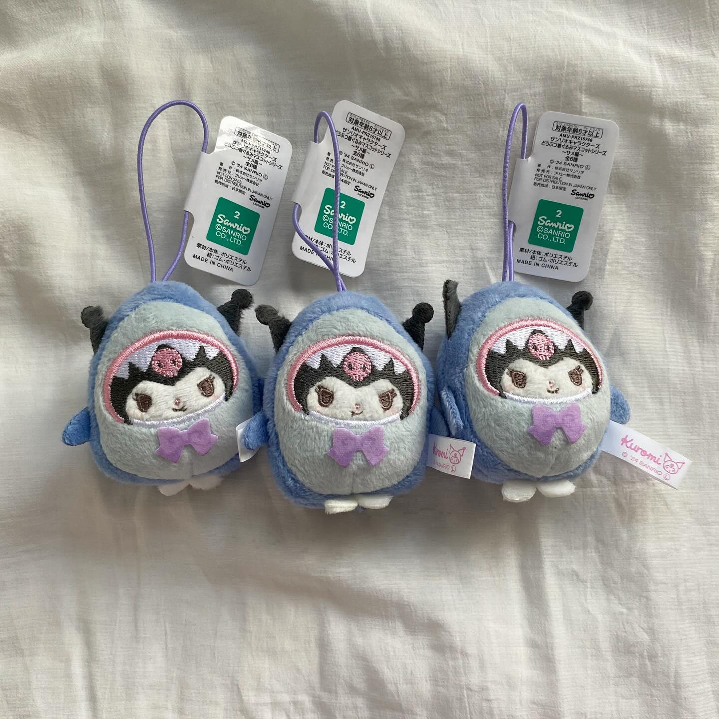 the image shows three kuromi plush keychains on a white fabric background. the keychains are designed as kuromi characters dressed in shark costumes. they have light blue shark bodies, gray inner faces with embroidered details, and light purple bows. each keychain is attached to a purple loop and has a white tag with sanrio branding and japanese text. the lighting is soft, and the overall aesthetic is cute and appealing to fans of kawaii culture and sanrio characters.