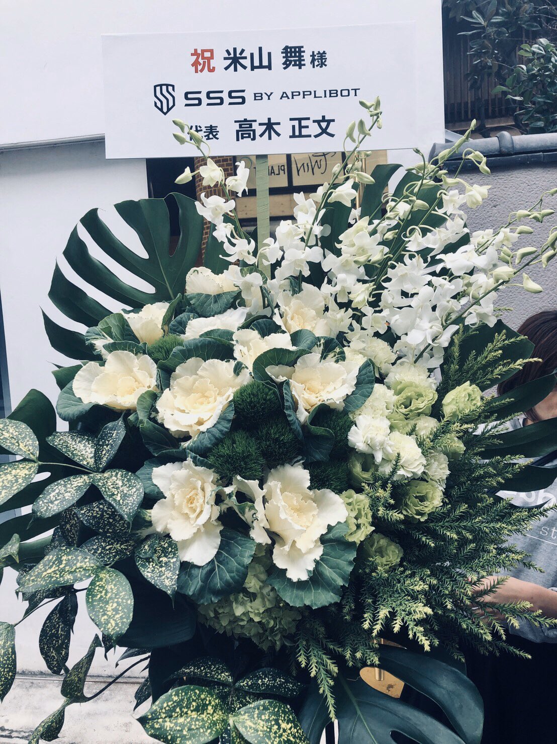 a large floral arrangement stands in front of a white wall with a sign on it. the sign, written in japanese, appears to celebrate the grand opening of a business. the flowers are primarily white and green, including orchids, monstera leaves, and other greenery. the overall atmosphere suggests a joyous and celebratory occasion.