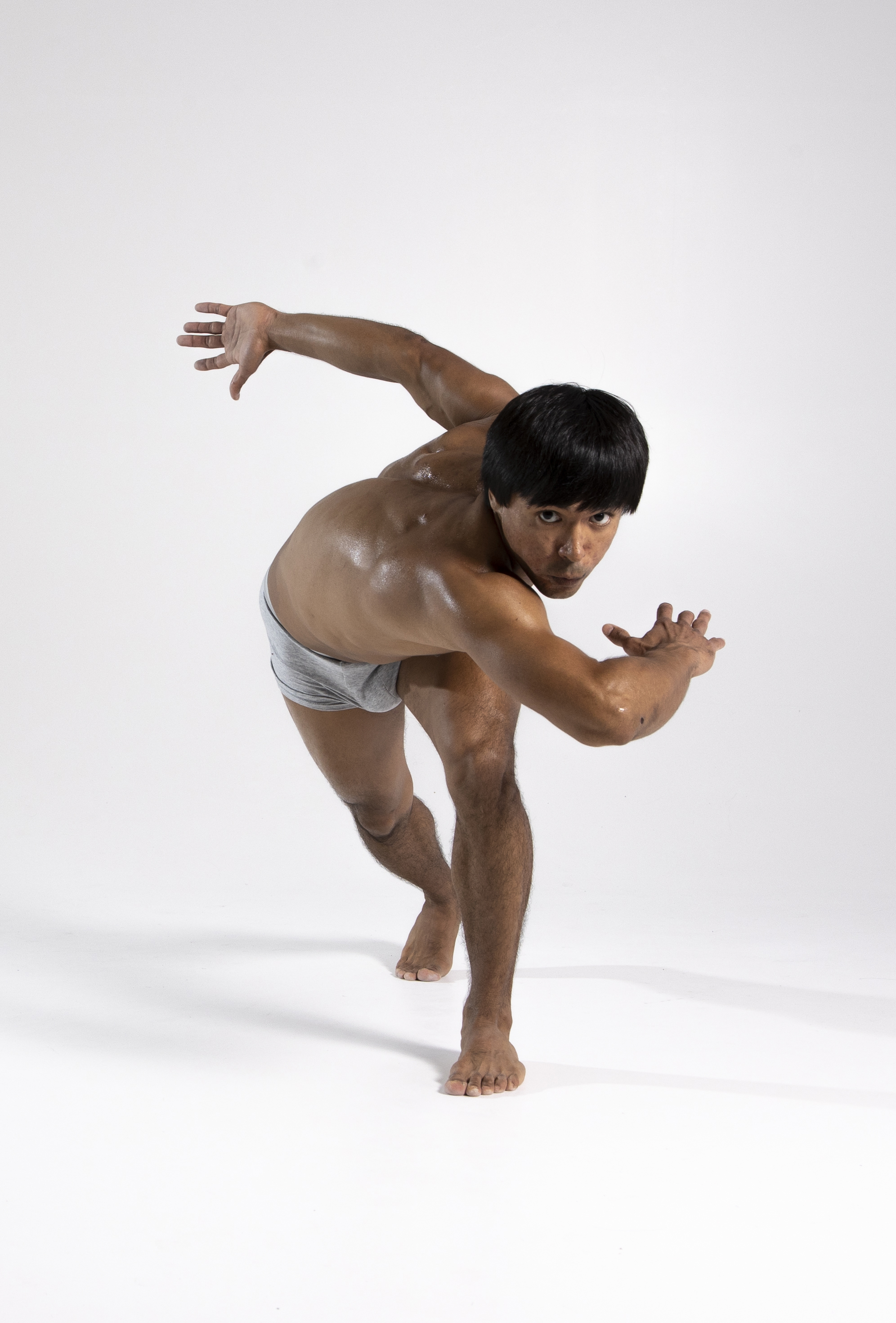 a muscular man with a bowl cut haircut is posing barefoot in a semicrouched, athletic stance. he is wearing shorts and standing indoors against a white background. his skin is glistening, possibly with sweat or oil. he is holding his arms out in front of him with his hands open.