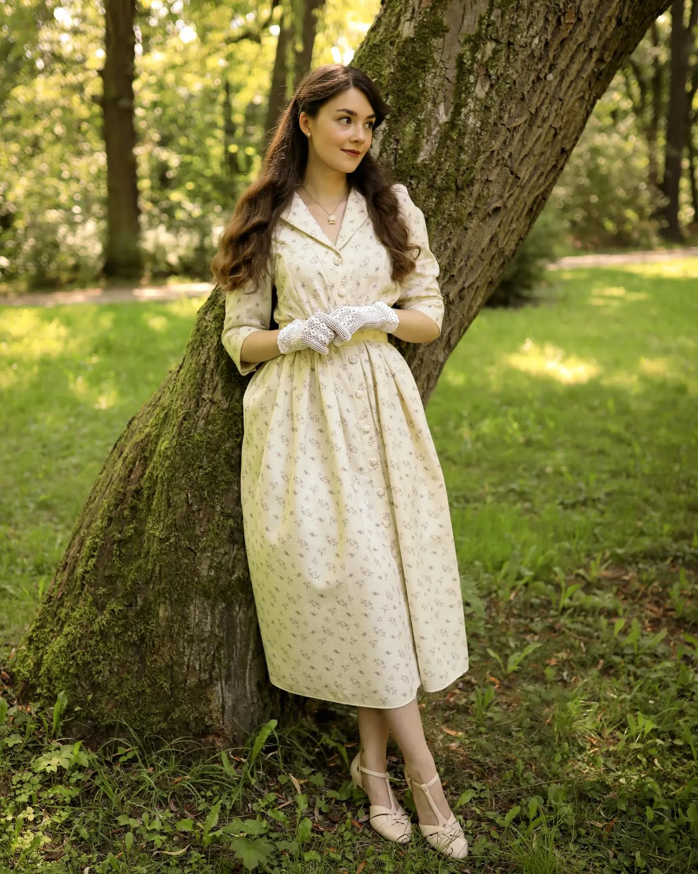 a young woman in a vintagestyle creamcolored midi dress with a delicate floral pattern stands gracefully beside a large tree in a park. the dress has threequarter sleeves and a buttondown front. she is wearing white lace gloves and beige heels.  her brown hair is styled in a soft wave. the setting is sunny and idyllic, suggesting a summertime scene.  the overall aesthetic is reminiscent of 1940s or 1950s fashion. the image is a fulllength portrait, focusing on the woman and her attire within the natural setting. she appears serene and happy.