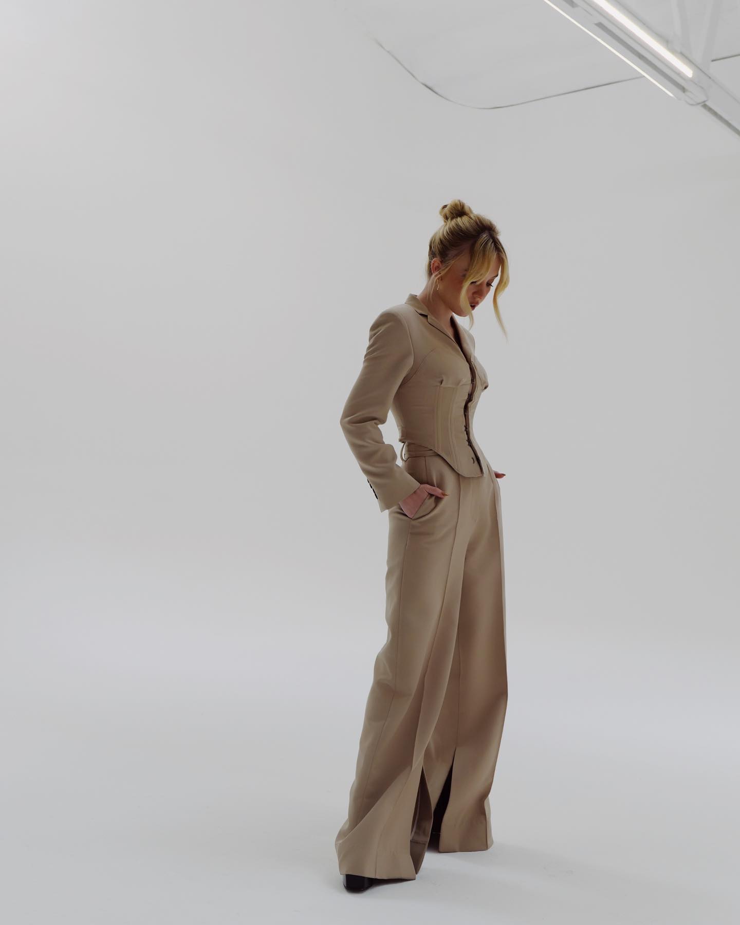 a fulllength shot of a blonde woman wearing a beige pantsuit. she is standing in a minimalist white studio, with her hands in her pockets, looking down. the suit is a highwaisted design with wideleg pants that have a slit on each side. the woman has her hair styled in a bun and is wearing minimal accessories. the overall aesthetic is elegant and sophisticated.