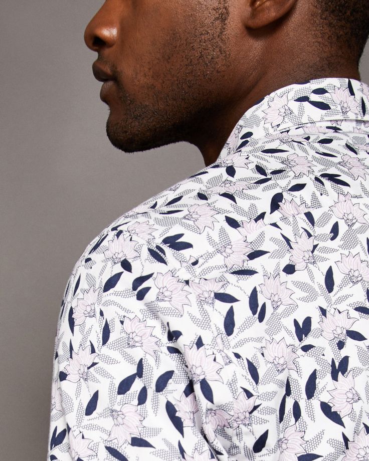 loseup side view of a man's shoulder and upper back, showcasing a white longsleeved shirt with a delicate floral print. the pattern features light pink flowers and navy blue leaves on a white background with a subtle textured pattern. the shirt is buttondown, and the collar is visible. the man's profile shows a dark complexion and short beard. the background is a muted gray, providing a neutral backdrop for the clothing detail.  the image focuses on the texture and pattern of the shirt fabric.