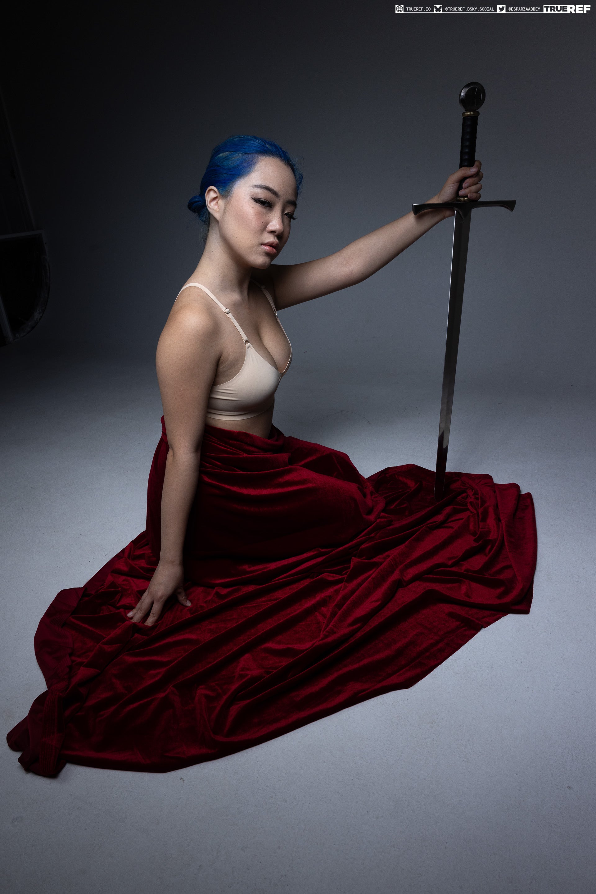 a woman with blue hair is sitting on the floor draped in red velvet, holding a sword in a studio setting. she is wearing lingerie and looking directly at the camera with a serious expression. the background is grey, and the lighting highlights the contrast between the velvet, her skin, and the metal of the sword. the trueref logo is visible in the upper right corner.