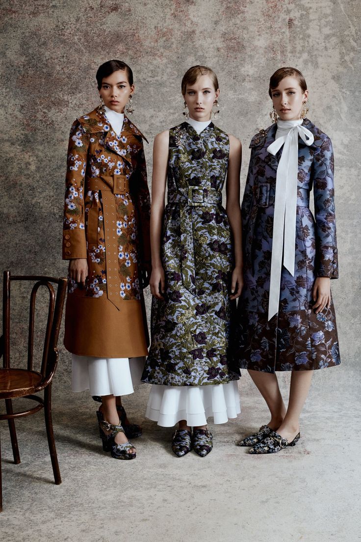 three female models are elegantly posed in a studio shot, showcasing a collection of autumn/winter attire. the model on the left wears a long floralprinted brown coat with a white midi skirt, the model in the center wears a sleeveless floral brocade midi dress, also with a white underskirt and ruffled hem, and the model on the right sports a longsleeved floral brocade coat in shades of blue and brown with a white bow detail.  all three models are wearing highheeled shoes with matching floral embroidery. the background features a textured grey wall. the overall atmosphere is sophisticated and fashionforward.