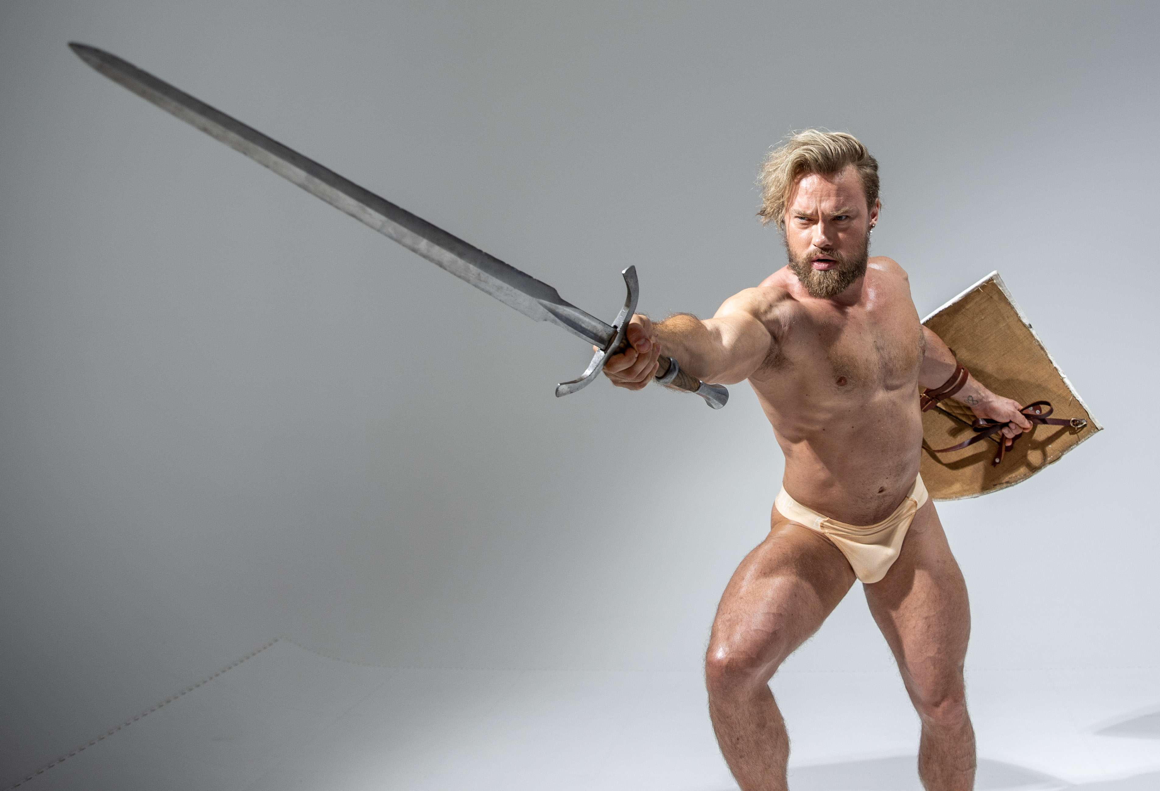 a muscular man with blonde hair and a beard is posed in a warrior stance against a light grey background. he is wearing a loincloth and wielding a sword and shield. the image is brightly lit, creating a heroic and powerful atmosphere.