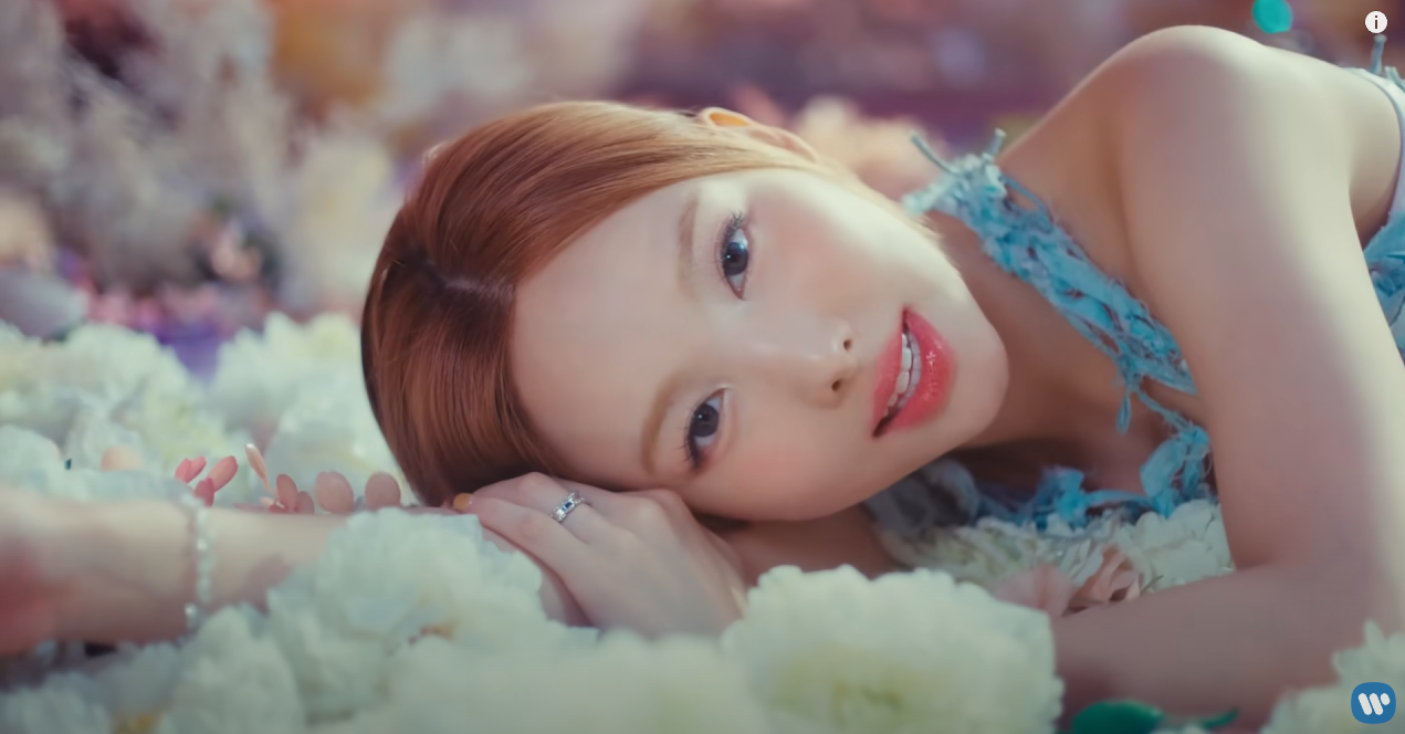 a young woman with long, reddishblonde hair lies on a bed of white flowers. she is wearing a light blue, ruffled top and delicate jewelry. her makeup is soft and romantic, with a focus on her eyes and lips. the image has a dreamy and ethereal quality, with soft lighting and a pastel color palette.