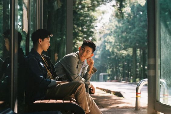 two young asian men are sitting on a bench at a bus stop, engaged in conversation. they appear happy and relaxed. the bus stop is located on a road that runs through a forest, with trees visible in the background. the scene is bathed in soft, natural light, creating a warm and inviting atmosphere.