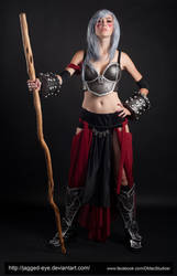 the image showcases a female cosplayer dressed as a fantasy warrior.  she's posed dramatically against a black backdrop, holding a wooden staff. her costume includes metallic armor, a dark, layered skirt (primarily black with burgundy accents), and sturdy boots with metallic details.  her hair is styled in a grey, shoulderlength fashion. the overall aesthetic is dark, powerful, and hints at a fantasy or game character portrayal. the lighting is dramatic, focusing on the cosplayer and her costume.