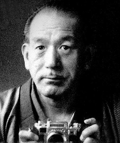 the image is a black and white closeup portrait of an older japanese man. he appears to be a photographer, holding a leicastyle rangefinder camera in front of him.  his expression is serious and contemplative. the background is dark and shadowy, drawing focus to the man's face and the camera. the overall mood of the photograph is artistic and slightly moody, suggesting a sense of experience and reflection. the image has a classic, vintage feel.