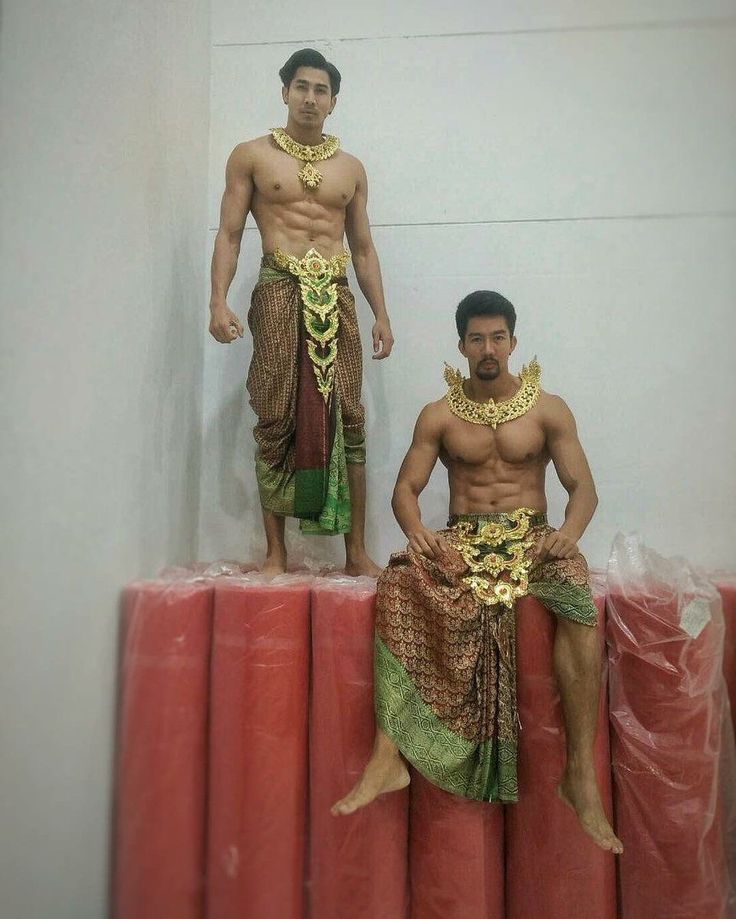 two men wearing traditional thai costumes with gold jewelry. one is standing and the other is sitting on a red background. they are both muscular and have a welltoned physique. they appear to be in a stage setting or backstage. the men are likely performers, actors, or dancers.