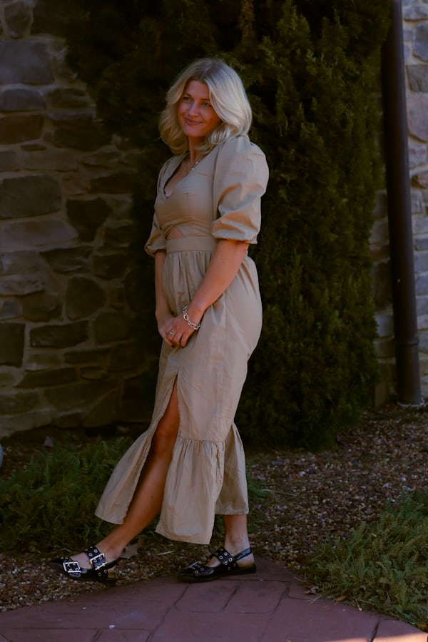a blonde woman in a beige midi dress with puff sleeves and a side slit stands outdoors against a stone wall.  the dress is a relaxed, casual style with ruffles at the bottom. she is wearing black studded sandals and is smiling slightly at the camera. the setting appears to be a garden or park area.