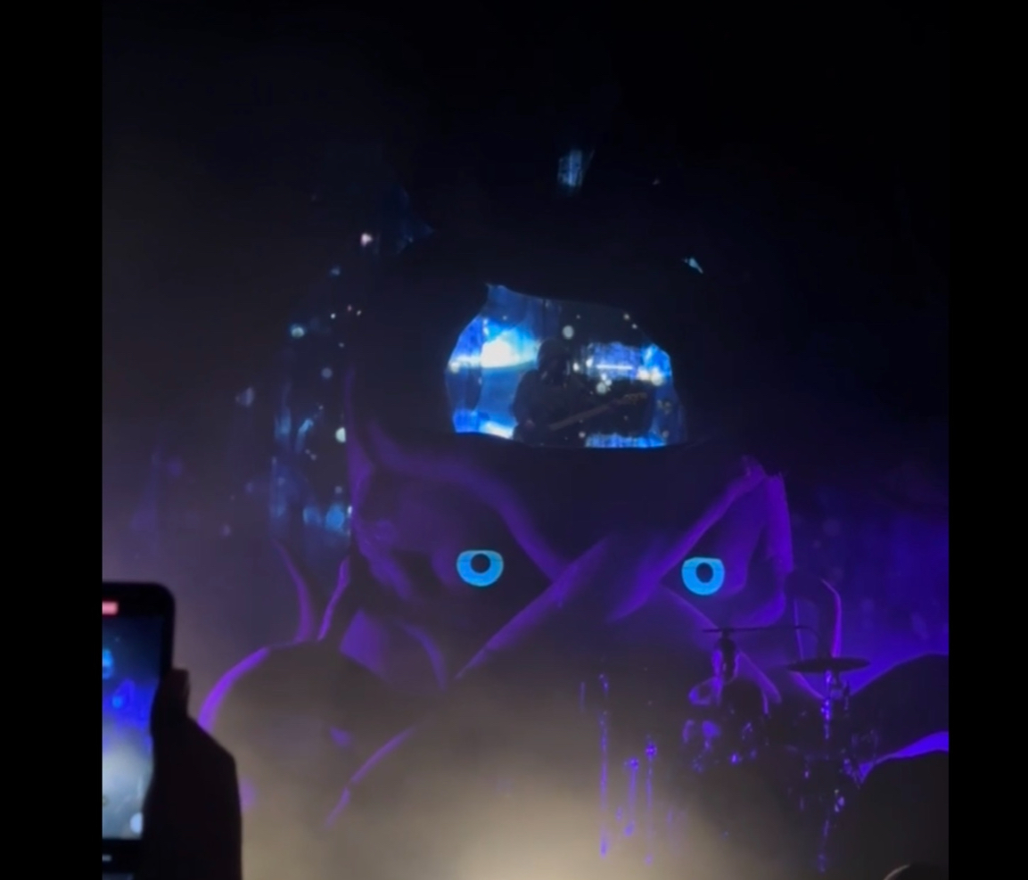 a dark, atmospheric photo of a concert stage. the stage is lit by blue and purple lights, and a large, mysterious figure is projected onto a screen. the figure has large, blue eyes and appears to be wearing a purple hood. the audience is silhouetted in the background, and a person in the foreground is holding up a phone to take a picture. the image evokes a sense of mystery and intrigue, and it is unclear what the figure is or what is happening on stage.