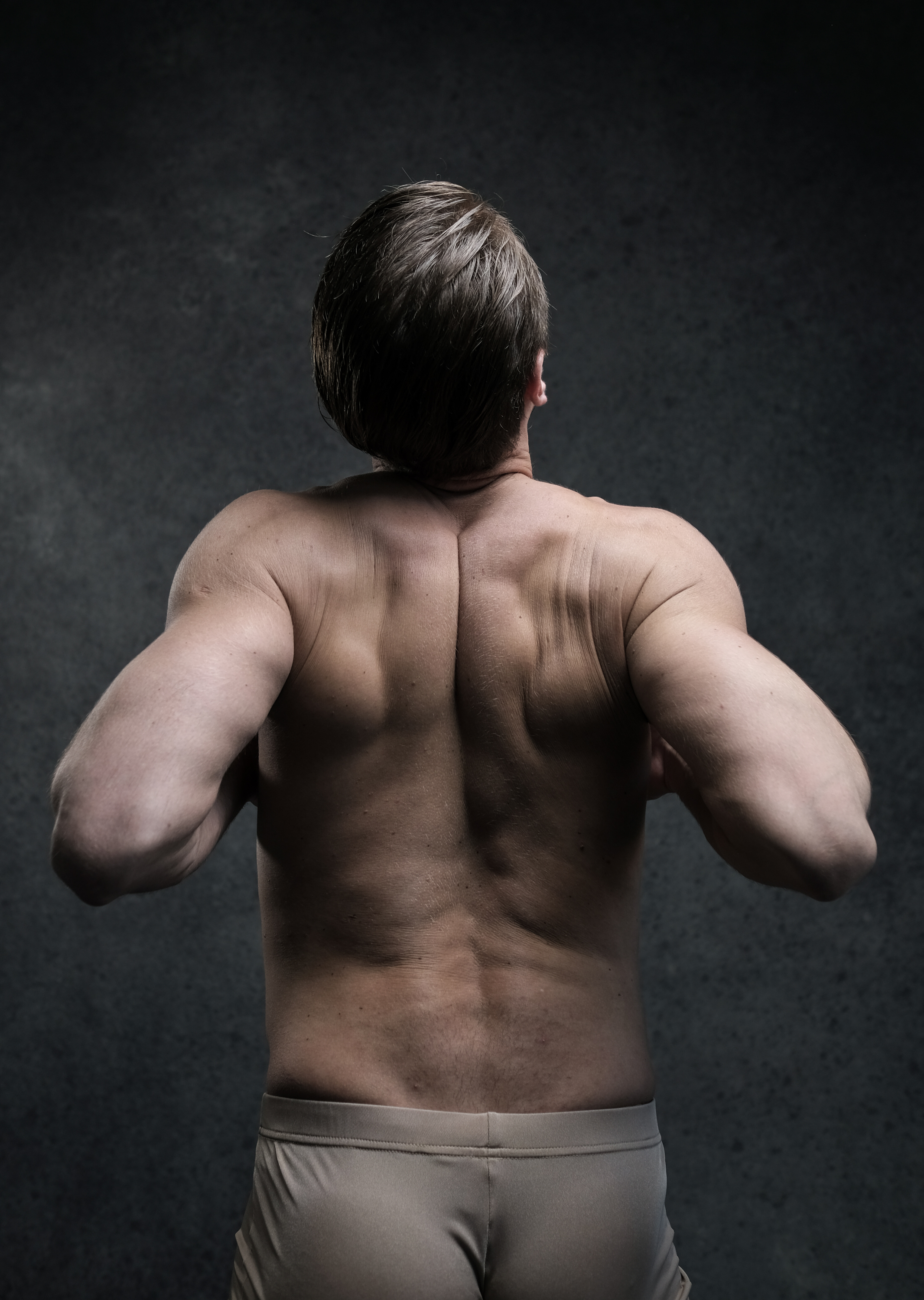 a rear view of a muscular man, illuminated by studio lighting, highlighting his toned back muscles. the man is standing with his arms extended, showcasing his physique.
