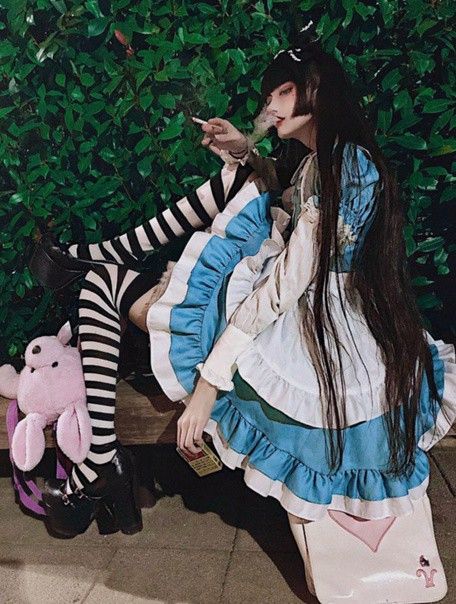 a young woman in a white and blue lolita dress with black and white striped stockings and black shoes sits on a bench with a cigarette in her hand. her long black hair covers her shoulders and falls to her waist. she is smoking the cigarette and looking away from the camera. the background is a wall of green plants. she is cosplaying as alice from alice in wonderland.  she is wearing a white frilly dress with a blue layer of ruffles underneath. her stockings are striped with black and white, and her shoes are black mary janes. she is holding a cigarette and her face is slightly smudged with what appears to be makeup. she is wearing a heartshaped necklace.