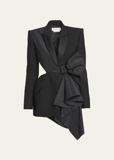the image shows a black blazer dress with an asymmetrical design. it features a large bow detail at the waist, a cutout on the side, and a tailored fit. the overall style is modern and formal, reflecting designer fashion for women.