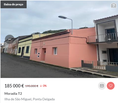a real estate sign in portuguese announcing a reduced price for a twostory house with a pink facade and a red roof. the house is located on a narrow street with a yellow house to the left and a house with white walls and a balcony to the right. the house is in good condition and has a streetlight in front of it. the sign shows the price of the house is 185,000 euros, which is 3% lower than the original price of 190,000 euros. the sign also mentions that the house is a twobedroom house and that it is located in the ponta delgada area of the island of são miguel in portugal.