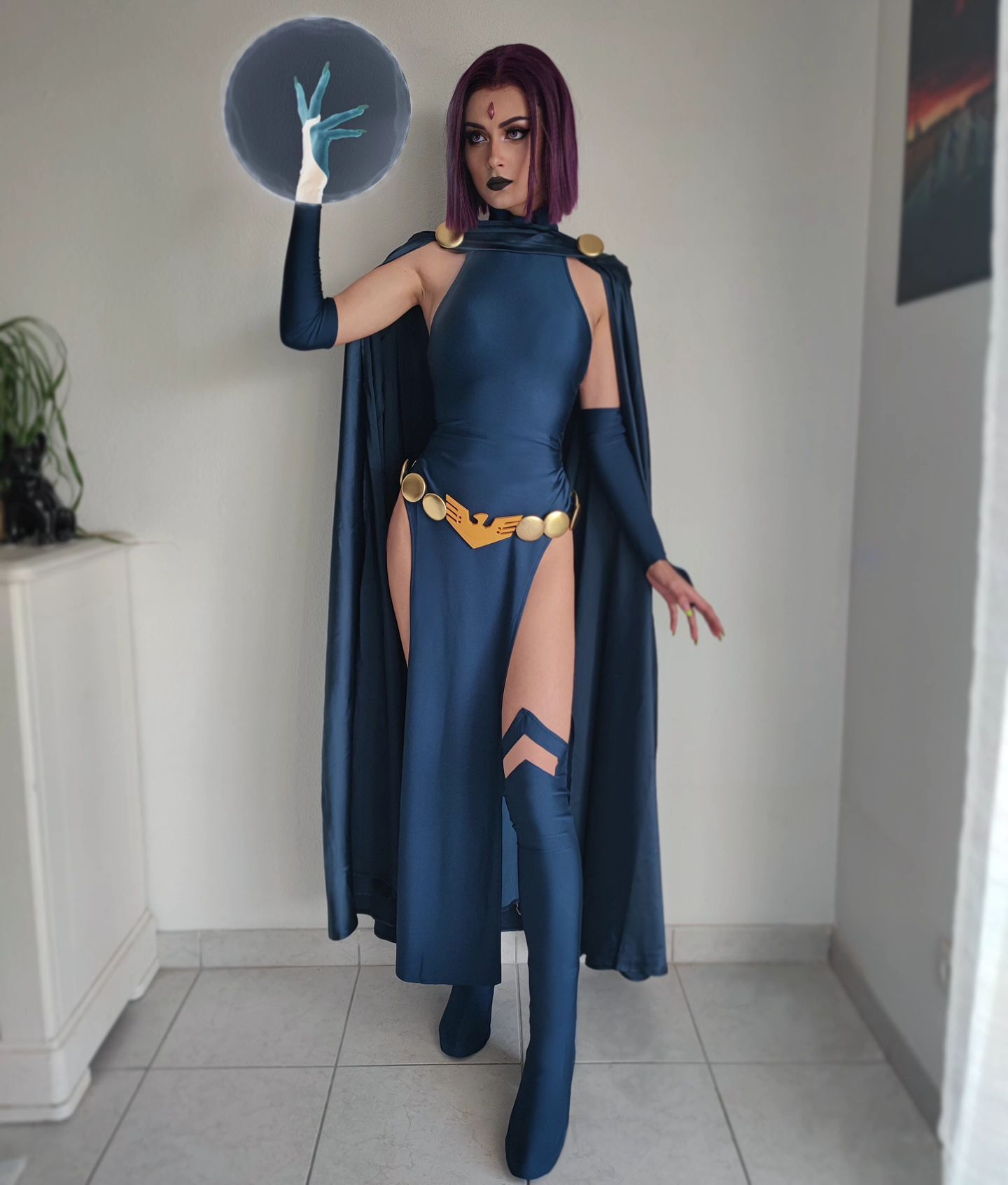 a fullbody shot of a woman cosplaying as raven from teen titans. she's wearing a dark blue dress with a cape, thighhigh boots, and a gold belt. her purple hair is styled in a bob. she is holding a glowing orb in her hand. the background is a plain white wall.