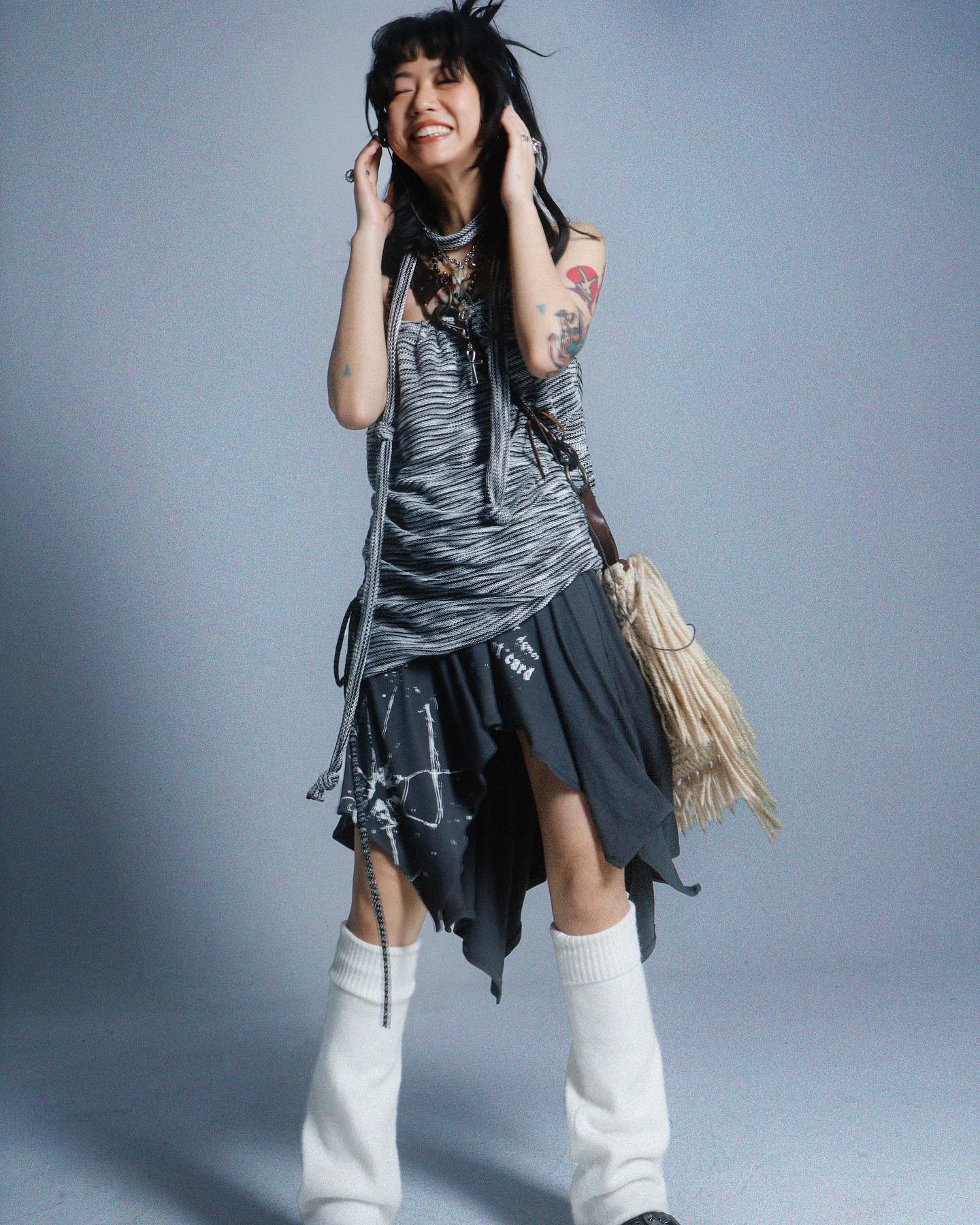 a young woman with dark hair and tattoos is smiling happily while listening to music with headphones. she is wearing a stylish and edgy outfit that includes a striped tank top, an asymmetrical darkgrey skirt with paint splatters, white socks, and a fringed bag. the overall style is alternative and grungeinspired. the photo is taken against a light grey background in a studio setting.