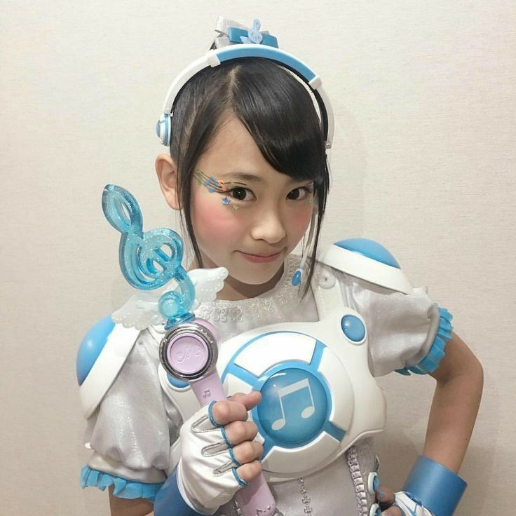 a young girl in a white and blue fantasy costume smiles at the camera. she is holding a sparkly blue musical note wand.  she has her dark hair pulled back and has glittering blue and rainbow makeup on her eyes.