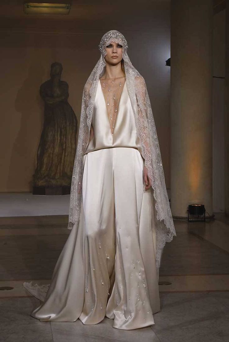 a fashion model walks the runway in a white wedding dress with a lace veil and a jeweled headpiece. she is posing for the camera, showing off the intricate details of her gown and the elegance of her look.  the dress features a plunging neckline, a flowing skirt, and a belt at the waist. the model's makeup is natural and understated, allowing the focus to remain on the gown. the overall look is sophisticated, glamorous, and chic.