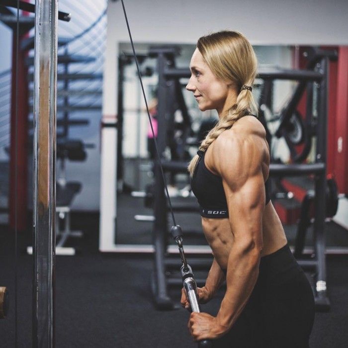 the image showcases a muscular woman intensely engaged in a triceps workout at the gym. she is positioned sideways, her back facing the camera, offering a clear view of her defined arm muscles as she pulls down on a cable machine. the setting is a typical gym environment with exercise equipment and mirrors lining the background, bathed in bright, artificial lighting.