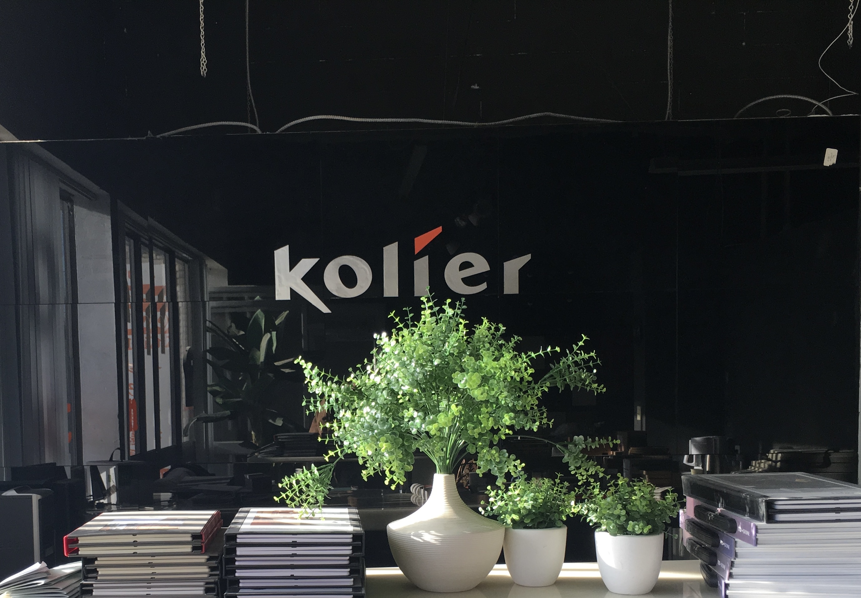 an interior shot of a store or office space. the background wall is black with a white "kolier" logo in the center.  there are three plants in white pots in front of the wall, one large plant in a large white vase, and two smaller plants in smaller white pots. in the foreground, there are two stacks of books. the overall aesthetic is modern and minimalist.