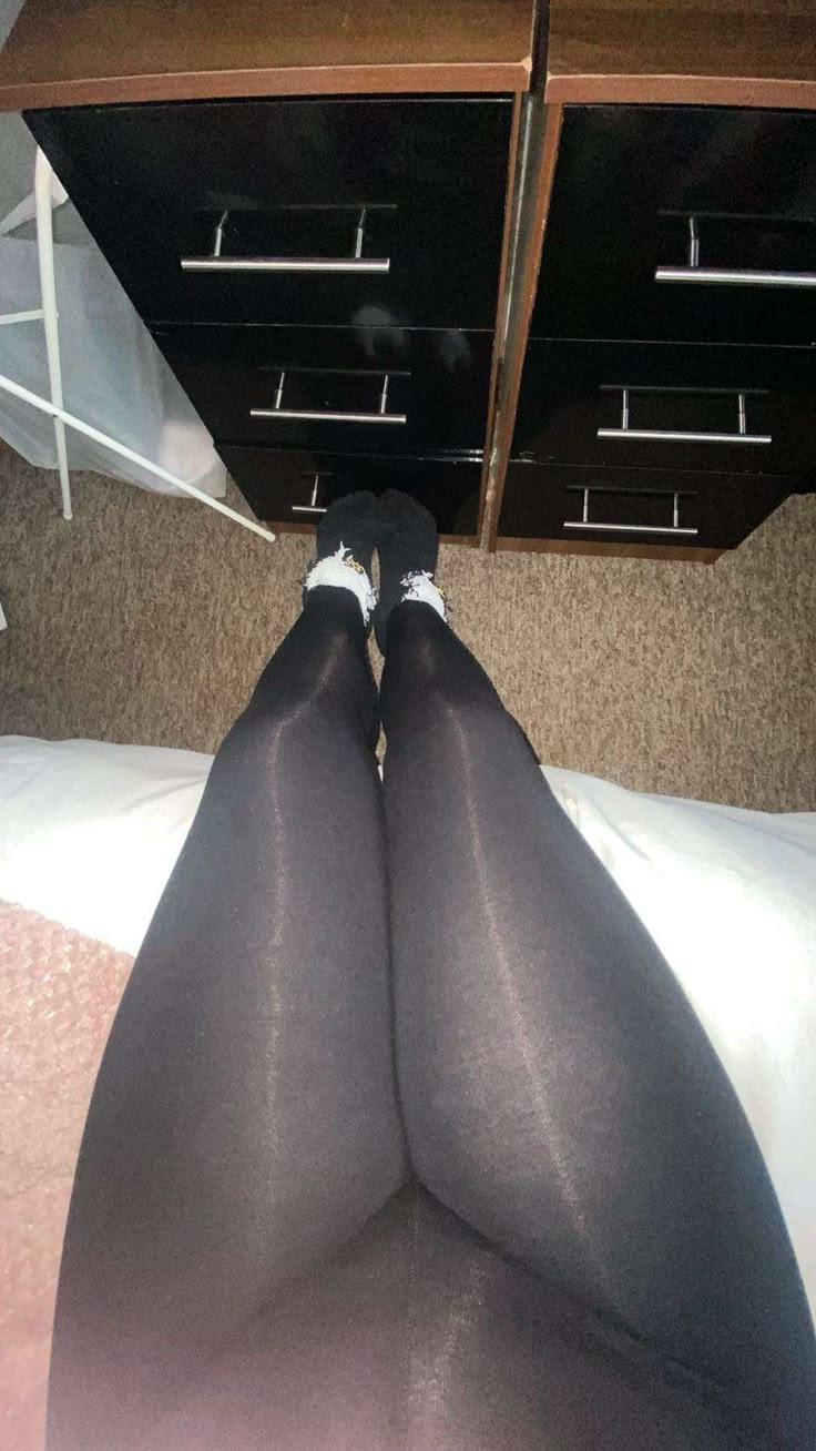 a view from above shows legs wearing black pantyhose and white socks, crossed on a bed with white sheets. behind the legs, a dresser with black drawers and brown top is visible. the legs are positioned against the background of the dresser. carpet flooring is partially visible around the bed and dresser.